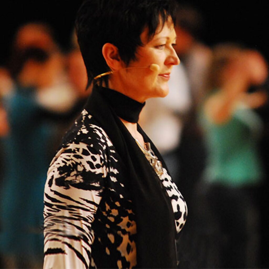 Martina Schürmeyer im Einsatz: Das Foto zeigt die Moderatorin in einer dynamischen Bühnensituation. Sie trägt ein stilvolles Outfit mit schwarz-weißem Muster und ist mit professioneller Audiotechnik (Headset) ausgestattet. Der unscharfe Hintergrund fängt die lebendige Atmosphäre einer Abendveranstaltung ein und rückt die Rednerin klar in den Fokus.